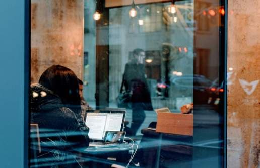 Zdjęcie zza szyby przedstawiające wnętrze restauracji i siędzącą osobę z laptopem